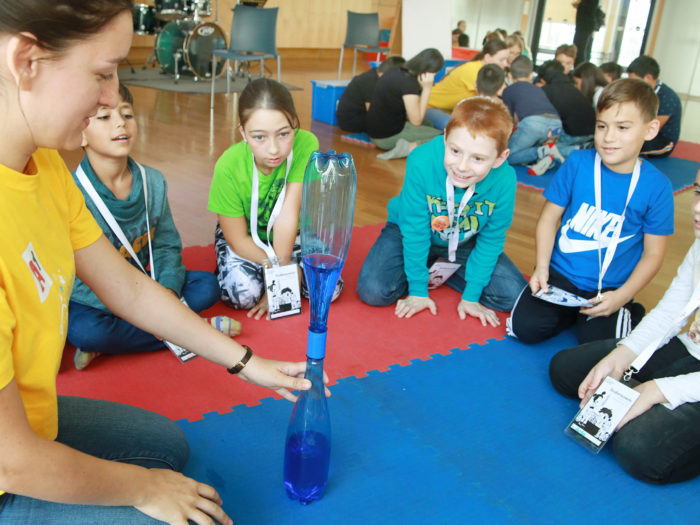 Kinder und Mitarbeiterin fürhen ein Experiment der Kinderuni on Tour mit zwei verbundenen Flaschen durch