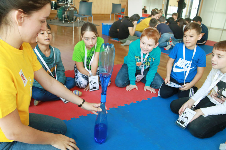 Kinder und Mitarbeiterin fürhen ein Experiment der Kinderuni on Tour mit zwei verbundenen Flaschen durch