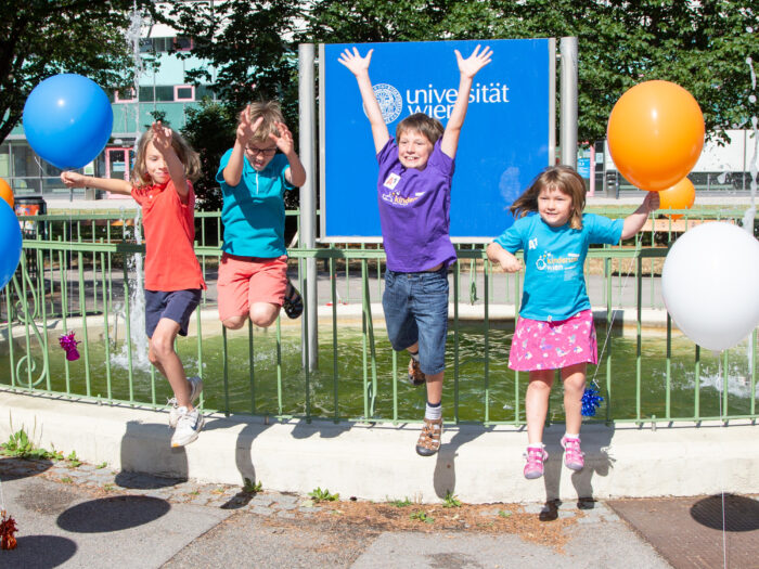 Kinder springen freudig in die Luft, dahinter der Springbrunnen mit dem Logo der Universität Wien, seitlich bunte Luftballons