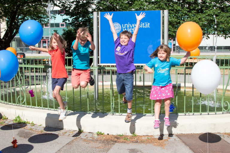 Kinder springen freudig in die Luft, dahinter der Springbrunnen mit dem Logo der Universität Wien, seitlich bunte Luftballons
