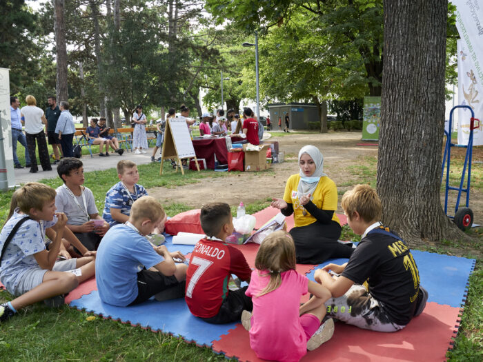 Kinder und ein Teammitglied sitzen im Park auf Matten und experimentieren