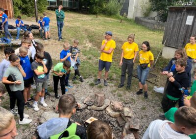Children and facilitators stand outside around a campfire site.